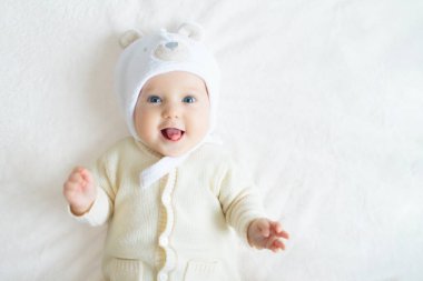 Beyaz güneşli yatak odasında şapkalı sevimli bir bebek. Yeni doğmuş çocuk yatakta dinleniyor. Küçük çocuklar için çocuk odası. Çocuklar için kış giysileri. Aile sabahı evde. Oyuncak ayıyla karnını doyururken doğan yeni çocuk..