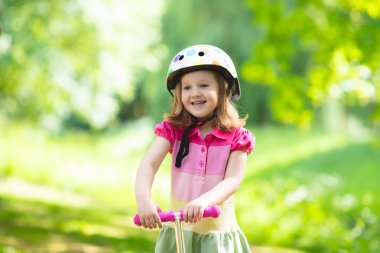 Küçük çocuk bir scooter bir şehir parkı güneşli yaz gününde binmek için öğrenme. Bir rulo sürme güvenlik kask şirin iskambil kız. Çocuklar açık havada oynar. Etkin boş ve çocuklar için açık spor.