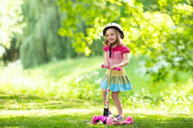 Küçük çocuk bir scooter bir şehir parkı güneşli yaz gününde binmek için öğrenme. Bir rulo sürme güvenlik kask şirin iskambil kız. Çocuklar açık havada oynar. Etkin boş ve çocuklar için açık spor.
