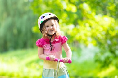Küçük çocuk bir scooter bir şehir parkı güneşli yaz gününde binmek için öğrenme. Bir rulo sürme güvenlik kask şirin iskambil kız. Çocuklar açık havada oynar. Etkin boş ve çocuklar için açık spor.