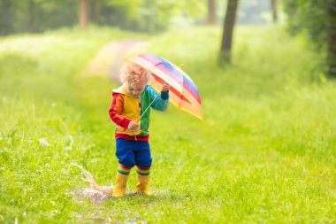 Yağmurlu yaz parkta oynarken küçük çocuk. Çocuk ile renkli gökkuşağı şemsiye, ceket ve su birikintisine atlama bot su geçirmez ve yağmurda çamur. Sonbahar duş yürüyüş çocuk. Herhangi bir weathe açık eğlence