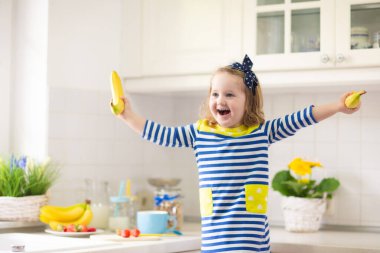 Çocuklar beyaz mutfakta kahvaltı yaparlar. Çocuklar için sağlıklı yiyecekler. Çocuk süt içiyor ve meyve yiyor. Mutlu gülümseyen anaokulu çocuğu sabah yemeğinin, mısır gevreğinin, muzun ve çileğin tadını çıkarıyor..