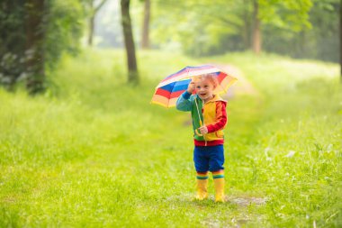 Yağmurlu yaz parkta oynarken küçük çocuk. Çocuk ile renkli gökkuşağı şemsiye, ceket ve su birikintisine atlama bot su geçirmez ve yağmurda çamur. Sonbahar duş yürüyüş çocuk. Herhangi bir weathe açık eğlence