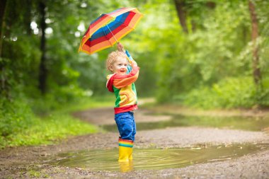 Yağmurlu yaz parkta oynarken küçük çocuk. Çocuk ile renkli gökkuşağı şemsiye, ceket ve su birikintisine atlama bot su geçirmez ve yağmurda çamur. Sonbahar duş yürüyüş çocuk. Herhangi bir weathe açık eğlence