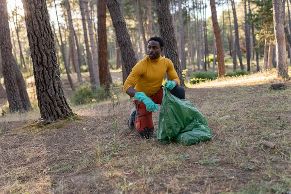 Young african american man collecting garbage into the plastic bag in the woods. People and ecology concept