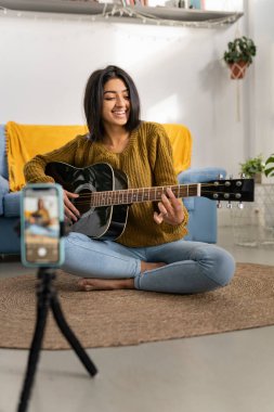 Genç esmer Hintli kadın akustik gitar çalarken akıllı telefonuyla kendi videosunu çekiyor. Milenyum Hindu dişisi evde.
