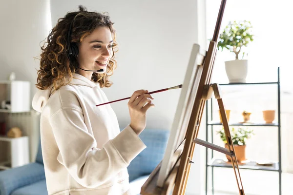 Happy young woman with curly hair painting a picture with headphones with microphone