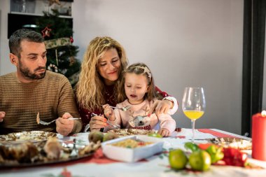 Baba ve anne ve kızları Noel yemeğini kutlamak için masada oturuyorlar. Annesi küçük kızını besliyor..