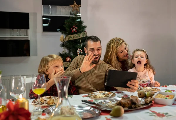 Caucasian family video calling from tablet during Christmas dinner. Father, mother, son and daughter waving