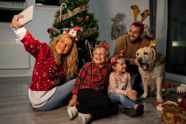 Kafkasyalı, noel bantlı ve labrador köpekli bir aile Noel ağacının yanında akıllı telefonuyla selfie çekiyor.