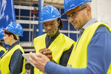Güvenlik kaskı ve yansıtıcı yelekli iki çok ırklı işçi tablete işçi eylemleri yazıyor. Bir robot fabrikasında. Afrika kökenli Amerikalı adama odaklan.