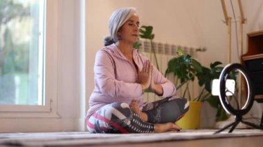 Yaşlı kadın evde yoga yapıyor. Dersi internetten kaydediyor.