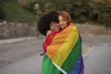 İki çok ırklı lezbiyen sokakta lgbtiq gökkuşağı bayrağıyla sarılıyorlar.