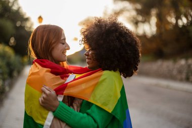 Çok ırklı lezbiyen çift gün batımında sokakta Igbt gökkuşağı bayrağıyla sarılarak birbirlerine bakıyorlar.