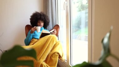 Beautiful young black woman sitting on the sofa reading a book and drinking coffee.