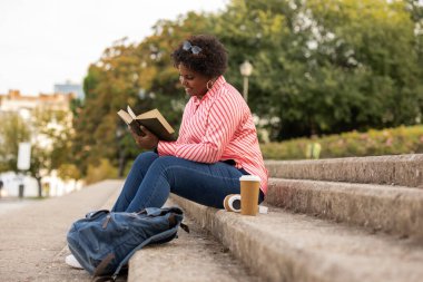 african american curvy woman student reading on stairs