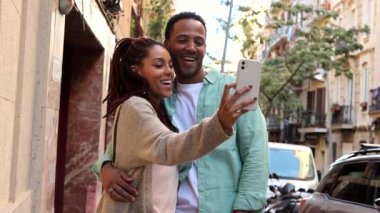 romantic relationship. Young diverse couple walking on city street embracing woman browsing internet on smartphone smiling joyful selfie.