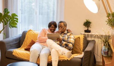 Smiling senior couple sitting on sofa at home making video call on digital tablet - African american married couple talk with family by video conference -