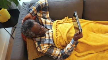 Handsome senior man lying on a sofa at home using a handheld tablet to buy gifts for friends and family online or make online video calls