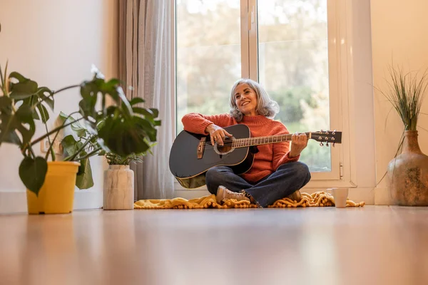 Smiling woman playing guitar - Mature woman with gray hair having online guitar lessons at home -