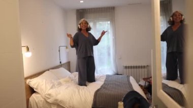 Exuberant mature woman jumping on the bed - listening to music and dancing at home happily -