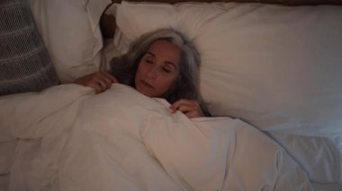 An old gray-haired woman lying on a pillow in bed woke up at night looking at the ceiling and tries to fall asleep. Insomnia sleep disorder, bad thoughts and anxiety, necessary treatment concept