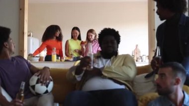 Çok ırklı bir arkadaş grubu televizyonda futbol maçı izlerken birayla kadeh kaldırıyor. Üç kadına odaklan.