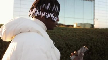 black man with dreadlocks walking through the city at sunset with a mobile phone