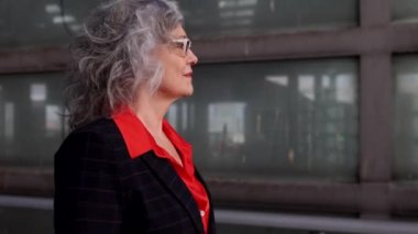 closeup of pretty senior woman with glasses and white hair, she is wearing formal clothes with a jacket, she is walking in the city at sunset