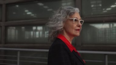 pretty senior woman with glasses and white hair, wears formal clothes with jacket, walks in the city at sunset