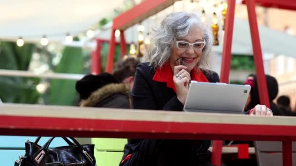 咖啡店里的女人在喝咖啡 在咖啡店里做数码平板电脑的女人 快乐的老年女性使用耳机 与家人或朋友联系 — 图库视频影像