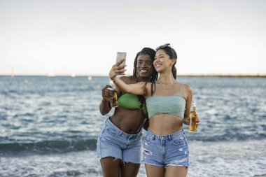 two multiracial female friends on the beach take selfie with smart phone, on the beach at sunset.