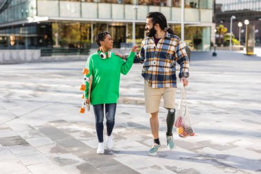 young couple on the street, multiracial, hispanic woman with short hair, caucasian man with a beard and a leg prosthesis, they walk and carry the purchase home