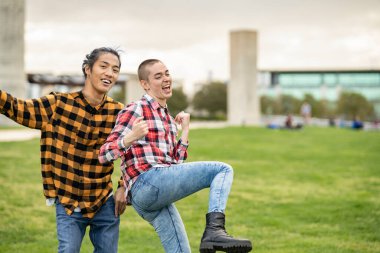 Two diverse people smiling and dancing outdoors- shaved Caucasian woman and long-haired Asian man, wearing casual clothes on campus.
