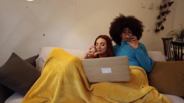 Evde dizüstü bilgisayarda film izleyen ve şarap içen iki kadın.