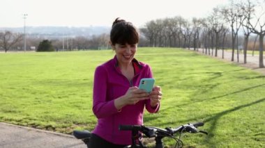 Dışarıda spor yapan, bisiklet ve kask takan oldukça olgun bir kadın akıllı telefonuyla telefon görüşmeleri için mola veriyor.