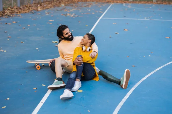 multiracial couple sitting skateboard, man has amputated leg and wears prosthesis.
