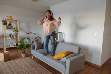 Oturma odası arka planında kablosuz kulaklıkla müzik dinlerken kanepede dans eden neşeli son sınıf öğrencisi kadın.
