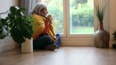 Gri saçlı olgun bir kadın, yerde battaniye ve bir fincan sıcak içecekle oturuyor, pencereden dışarı bakıyor.
