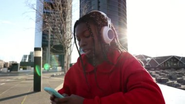 Genç Afrikalı bir kadın şehir binalarının önünde oturmuş akıllı telefonuyla müzik dinliyor.