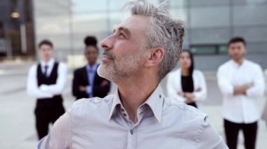 Orta yaşlı işadamı ofis binasının önünde kollarını kavuşturmuş duruyor. Dışarıda poz veren çok ırklı iş adamları.