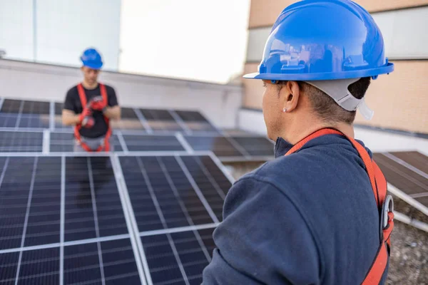 Male team workers in protective helmet installing solar photovoltaic panel system using screwdriver. Electrician mounting blue solar module on roof of modern house.