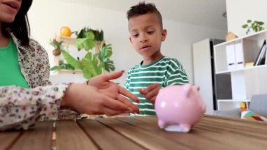 Annesiyle birlikte küçük Latin çocuk domuz kumbarasına para koyuyor. Tasarruf kavramı. -