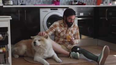 Sakallı adam mutfaktaki halıda köpeğinin yanında kahve içiyor.