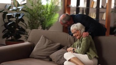 Gri saçlı büyükanne ve büyükbaba evde poz veriyor. Sevgi dolu eş, kanepede oturan kocaya sarılır, birlikte vakit geçirirler. Mutlu ve uzun süreli evlilik, eşler arasındaki uyumlu ilişkiler, saygı ve bağ.