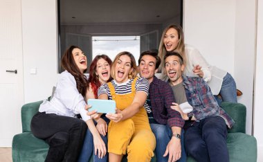 Bir grup farklı arkadaş evde kanepede eğleniyor, akıllı telefonlarıyla sosyal medya için selfie çekiyorlar.