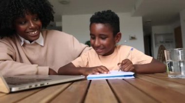 Anne oğlunun ödevlerine yardım ediyor, Afrikalı aile evde dizüstü bilgisayar ve defterle.