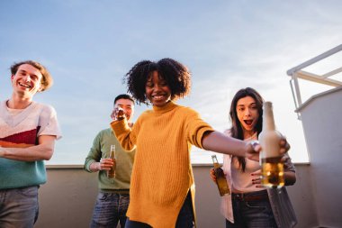 Çatıda birayla kadeh kaldırırken, dışarıda kutlama yapan çok ırklı arkadaşlar.