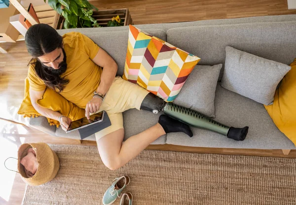 stock image Overhead view portrait of man with prosthetic leg doing homework lying on sofa looking at digital tablet