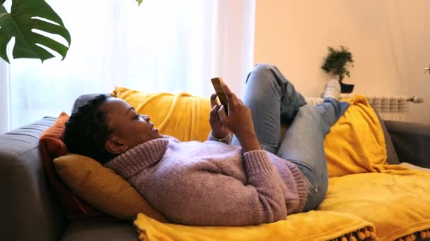 Африканская Молодая Женщина Лежит Диване Болтая Смартфоном Дома Социальные Сети — стоковое видео